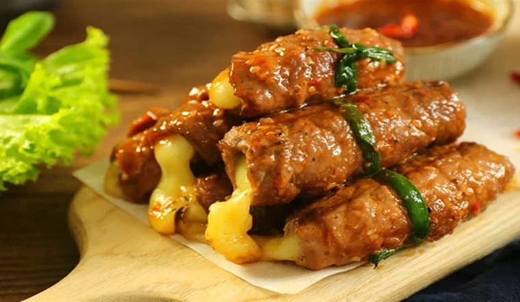 Món ba chỉ bò cuộn phô mai béo ngậy, thịt bò mềm thơm bao bọc nhân phô mai tan chảy, được buộc bằng hành lá và nướng vàng hấp dẫn, bày trên thớt gỗ.