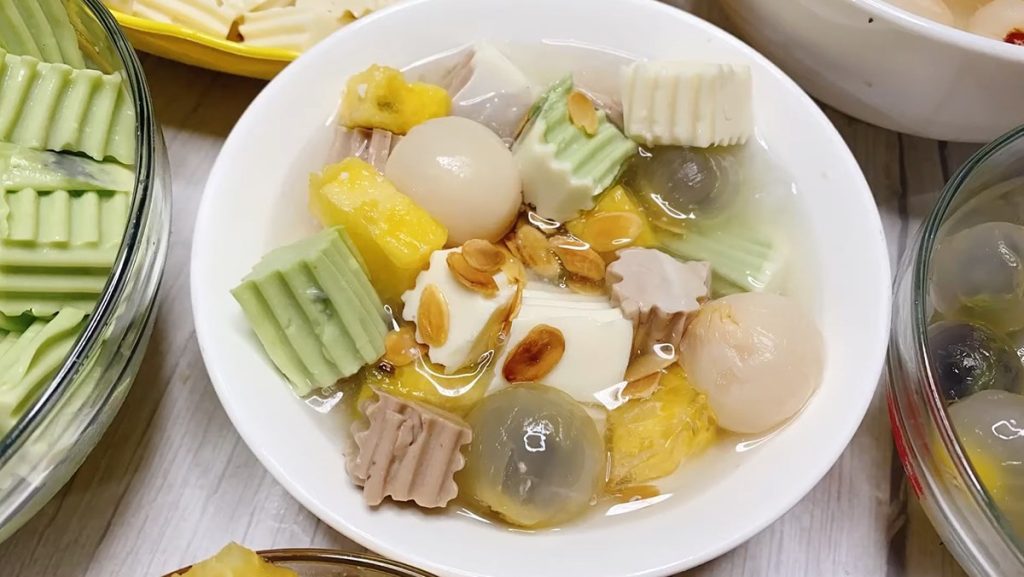 Chè khúc bạch hoa quả thanh mát với miếng khúc bạch nhiều màu, nhãn, dứa, thạch giòn và hạnh nhân rang, được bày trong bát trắng hấp dẫn.