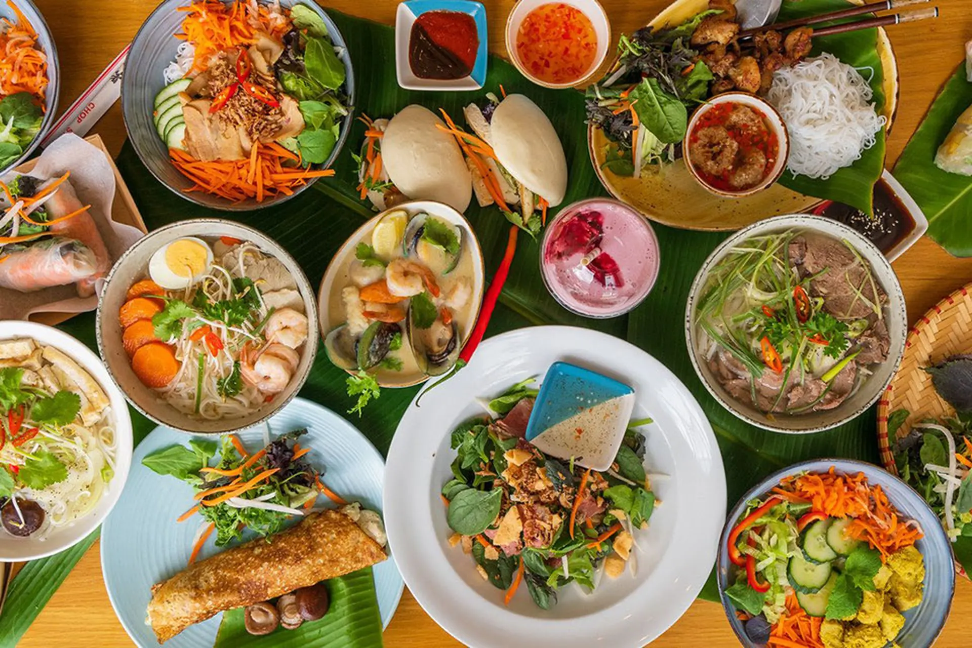Bàn tiệc ẩm thực Bắc Bộ với nhiều món ăn truyền thống và hiện đại như bún, phở, gỏi, nem rán, bánh bao kẹp, hải sản, rau xanh và nước chấm, được bày trí đẹp mắt trên lá chuối và đĩa sứ.