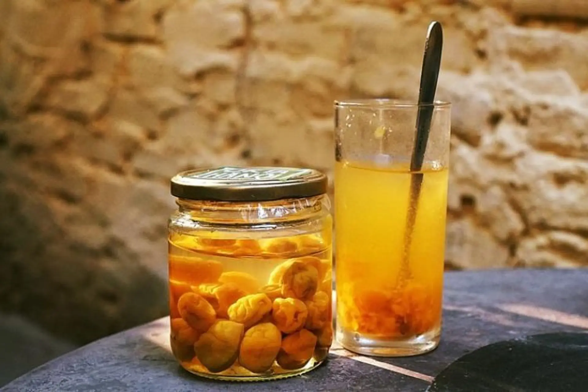 Lọ thủy tinh đựng mơ ngâm đường màu vàng nâu, có thể nhìn rõ những quả mơ tròn trịa, căng mọng bên trong, đặt cạnh một ly cao chứa nước mơ đã pha, có màu vàng cam nhạt. Chiếc thìa kim loại màu đen được đặt trong ly nước mơ. Bối cảnh phía sau là bức tường đá màu nâu nhạt.