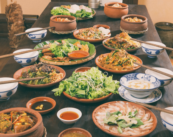 Một bàn tiệc dài bày trí theo phong cách ẩm thực Việt Nam truyền thống với nhiều bát gốm và đĩa đất nung. Các món ăn bao gồm rau sống, thịt luộc, đồ xào và cơm trắng, đi kèm với bát đũa sứ trắng hoa văn xanh cổ điển trên mặt bàn gỗ tối màu.