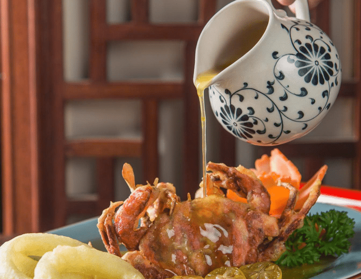 Ảnh cận cảnh nghệ thuật ghi lại khoảnh khắc rưới nước sốt vàng óng từ một bình sứ hoa văn xanh lên món cua lột chiên giòn. Phía sau là các vật dụng trang trí bằng gỗ và rau củ tỉa hoa, tạo cảm giác cao cấp và tinh tế.