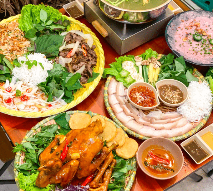 Một bàn tiệc thịnh soạn với tiêu điểm là gà nướng nguyên con vàng rộm, mẹt bún thịt luộc và các món cuốn. Các mẹt đồ ăn lớn được sắp xếp đan xen với bát nước chấm và một nồi lẩu đang sôi, thể hiện một bữa ăn cộng đồng phong phú.